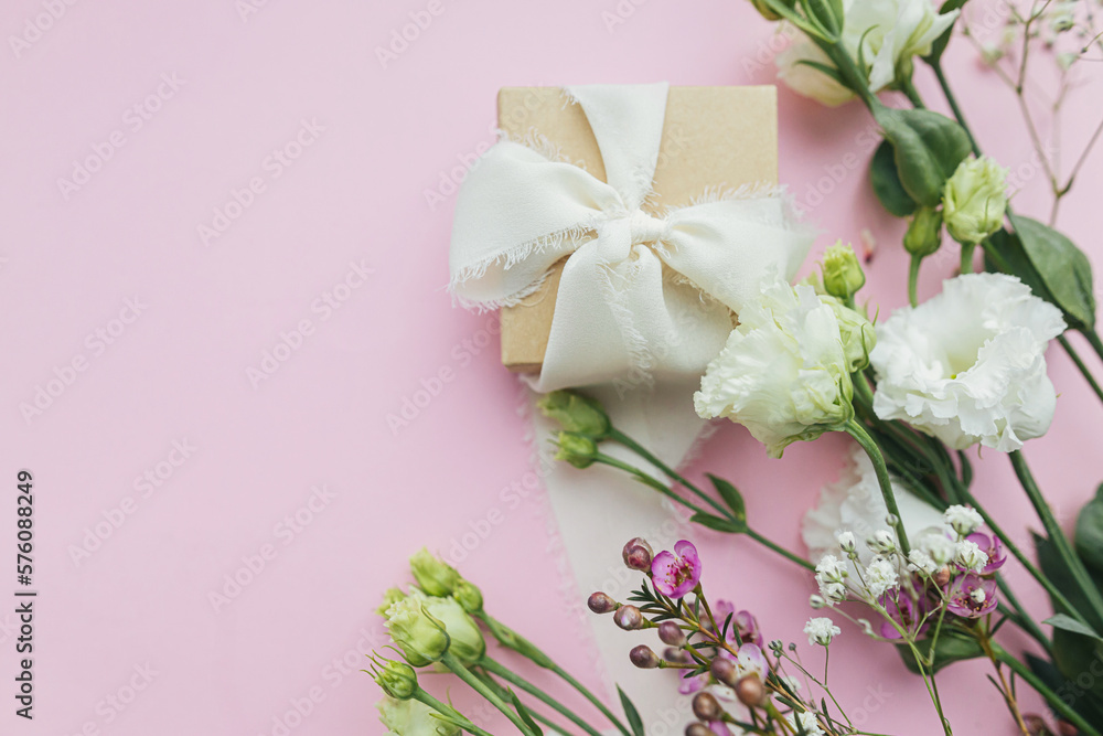 Stylish simple gift with ribbon and beautiful tender flowers on pink background flat lay with space for text. Happy womens day and mother's day concept. Floral greeting card or banner