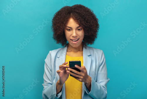 Excited African American woman holding mobile phone reads a unexpected message with good news standing on a blue background, emotional facial expression