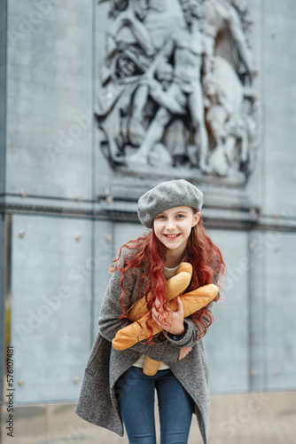 Pretty cheerful elegant young girl teenager with red hair in beret and coat with french baguettes leaning towards camera and laughing at city street, joyful outdoor emotional vertical urban portrait © upparadox