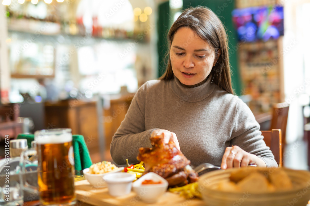 Smiling woman tourist enjoying traditional baked pork knuckle served with pickles, sauces and glass of beer in Viennese restaurant