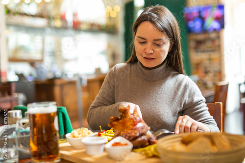 Smiling woman tourist enjoying traditional baked pork knuckle served with pickles  sauces and glass of beer in Viennese restaurant