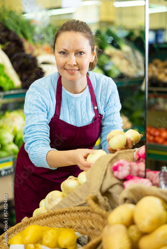 Positive middle-aged saleswoman setting out onions on food stall in big grocery store