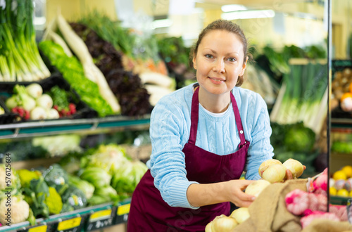 Skillful busy middle-aged female seller in apron putting fresh onion on shelves in grocery shop