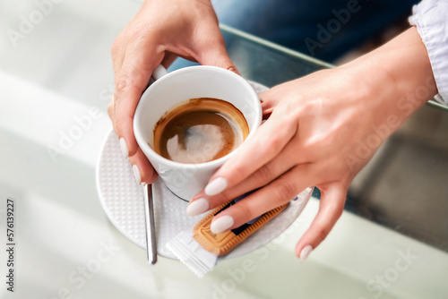 White cup with black coffee in the hands of a girl in a summer cafe. A girl in a white shirt and blue jeans holds a white cup of coffee in her hands with a manicure. Cup of coffee on a sunny morning