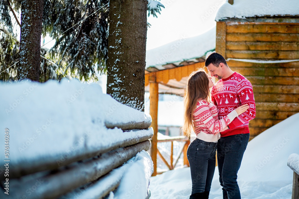 Engagement session, love story of a straight young couple hugging and kissing in Christmas red sweaters and jeans happily looking at each other in snowy landscape near a wooden house and pine trees