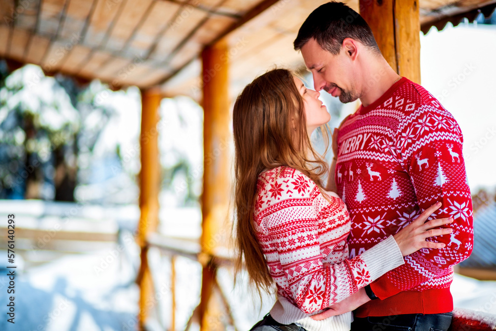 Engagement session, love story of a young straight couple hugging and kissing in Christmas red sweaters and jeans happily looking at each other at a wooden porch in a snowy mountain house
