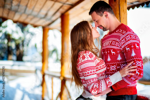 Engagement session, love story of a young straight couple hugging and kissing in Christmas red sweaters and jeans happily looking at each other at a wooden porch in a snowy mountain house © Michael Rekochinsky