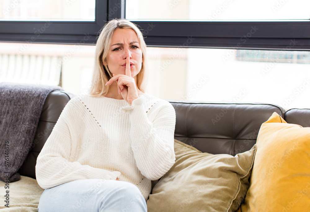 young pretty woman looking serious and cross with finger pressed to lips demanding silence or quiet, keeping a secret sitting on a couch young pretty woman looking serious and cross with finger pressed to lips demanding silence or quiet, keeping a secret sitting on a couch