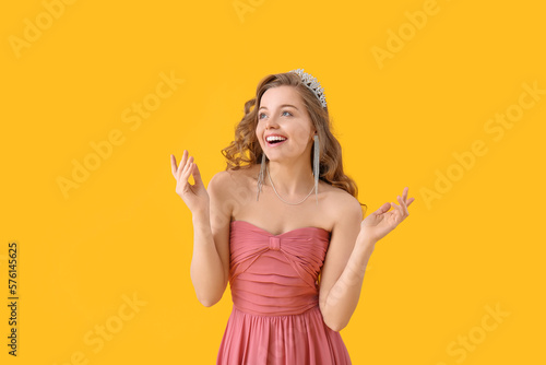 Happy young woman in tiara and prom dress on yellow background