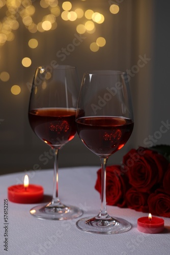 Glasses of red wine, rose flowers and burning candles on white table against blurred lights. Romantic atmosphere