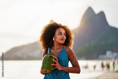 waist up portrait cheerful young brazilian afro hairstyle woman walking on the beach holding a coconut water in Ipanema 