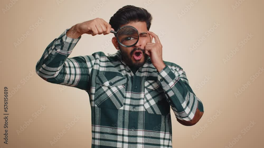 Investigator researcher scientist man holding magnifying glass near face, looking into camera with big zoomed funny eyes, searching, analysing. Handsome indian guy isolated on beige studio background Investigator researcher scientist man holding magnifying glass near face, looking into camera with big zoomed funny eyes, searching, analysing. Handsome indian guy isolated on beige studio background