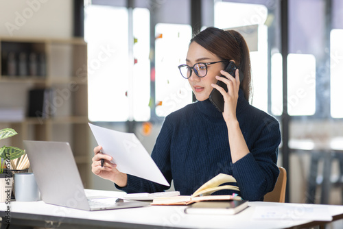 Cheerful business Asian woman freelancer making telephone call share good news about project working in office workplace, business finance concept.	