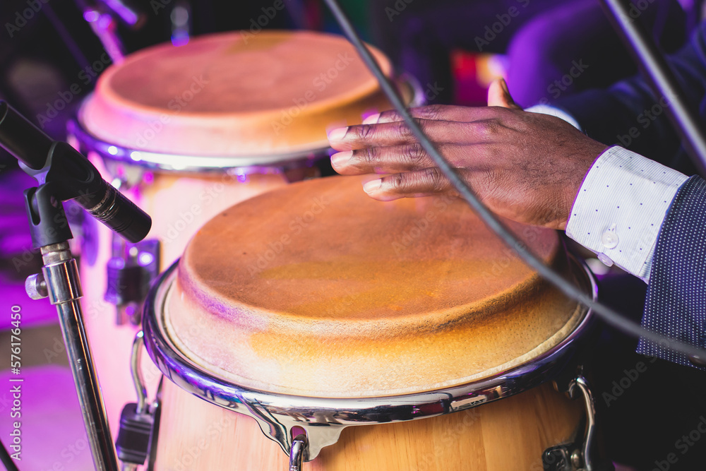 Fototapeta premium Bongo drummer percussionist performing on a stage with conga drums set kit during jazz rock show performance, tumbadora quinto with latin cuban afro-cuban jazz band performing in the background