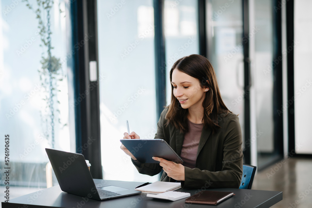 Confident business expert attractive smiling young woman typing laptop ang holding digital tablet  on desk in creative office.