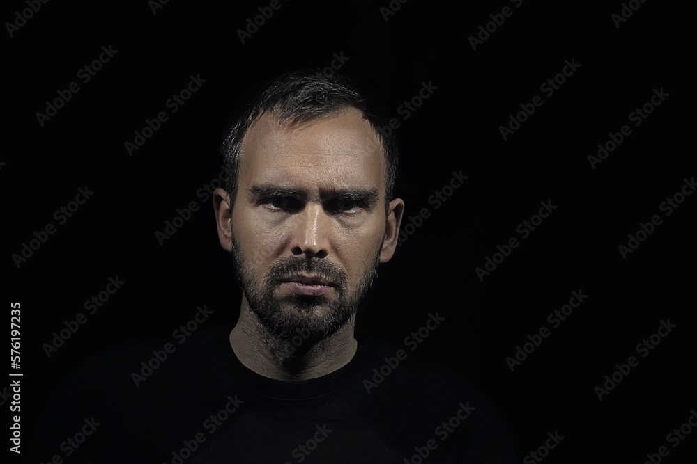 A serious man with grey hair and a small beard looks sullenly into the camera. A portrait of a Caucasian man with frowning eyebrows. A dramatic portrait on a dark background.