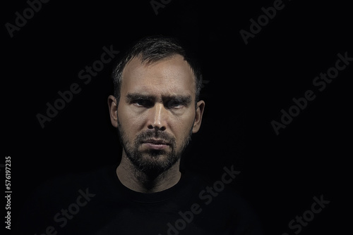 A serious man with grey hair and a small beard looks sullenly into the camera. A portrait of a Caucasian man with frowning eyebrows. A dramatic portrait on a dark background. photo