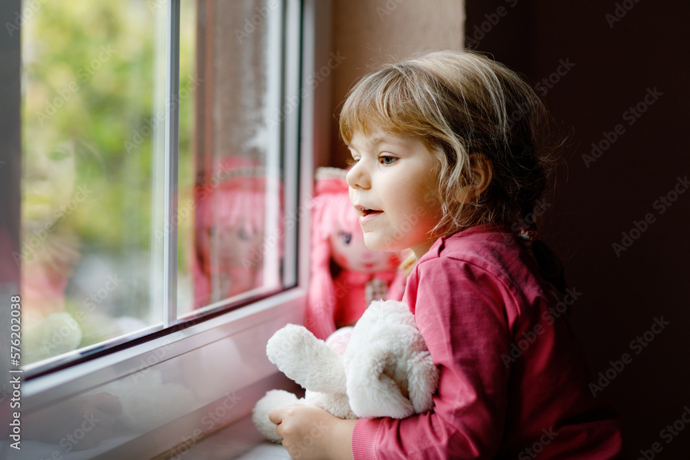 Cute toddler girl sitting by window and looking out on rainy day. Dreaming child with doll and soft toy feeling happy. Self isolation concept during corona virus pandemic time. Lonely kid. Cute toddler girl sitting by window and looking out on rainy day. Dreaming child with doll and soft toy feeling happy. Self isolation concept during corona virus pandemic time. Lonely kid.