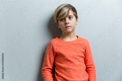 A sad boy of 9-10 years old stands near a gray wall. The guy in the orange sweater. Loneliness and depression.