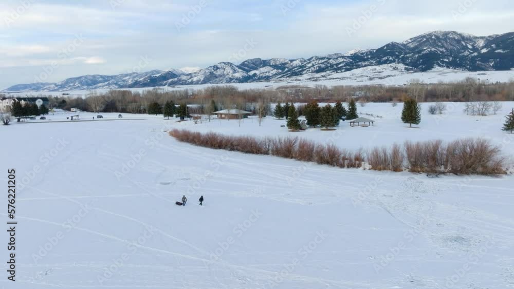 Boseman Montana Aerial Winter sunset ice fishermen pulling sled over snowy suburban park, orbit right with 4k drone with mountain backdrop at golden hour Boseman Montana Aerial Winter sunset ice fishermen pulling sled over snowy suburban park, orbit right with 4k drone with mountain backdrop at golden hour