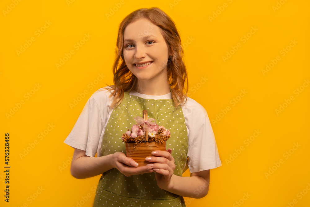 Happy smiling young woman in green polka dot apron and white shirt holding pink flowers in wicker basket and looking at camera isolated on yellow background. Floristry and floristic industry concept. Happy smiling young woman in green polka dot apron and white shirt holding pink flowers in wicker basket and looking at camera isolated on yellow background. Floristry and floristic industry concept.