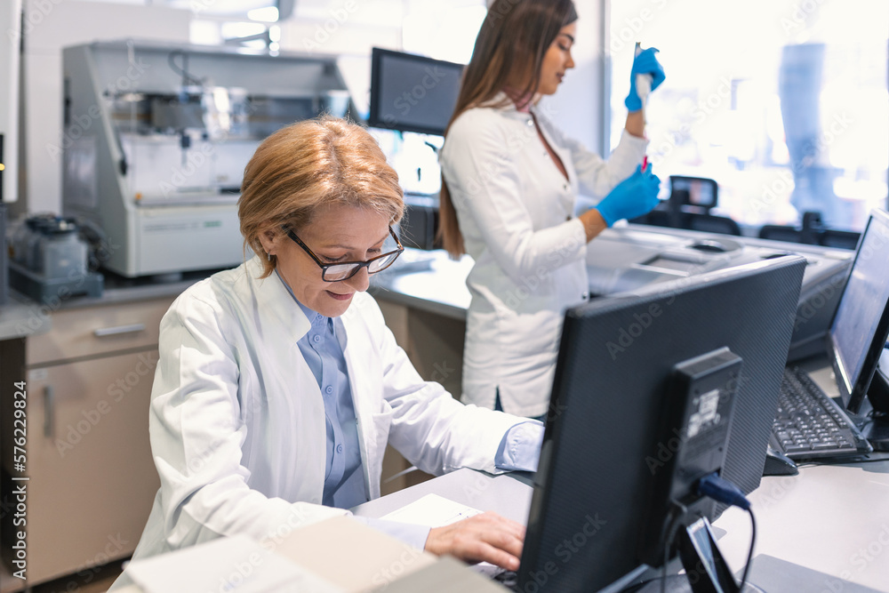 Female Scientists Working on their Computers In Big Modern Laboratory. Various Shelves with Beakers, Chemicals and Different Technical Equipment is Visible. Female Scientists Working on their Computers In Big Modern Laboratory. Various Shelves with Beakers, Chemicals and Different Technical Equipment is Visible.