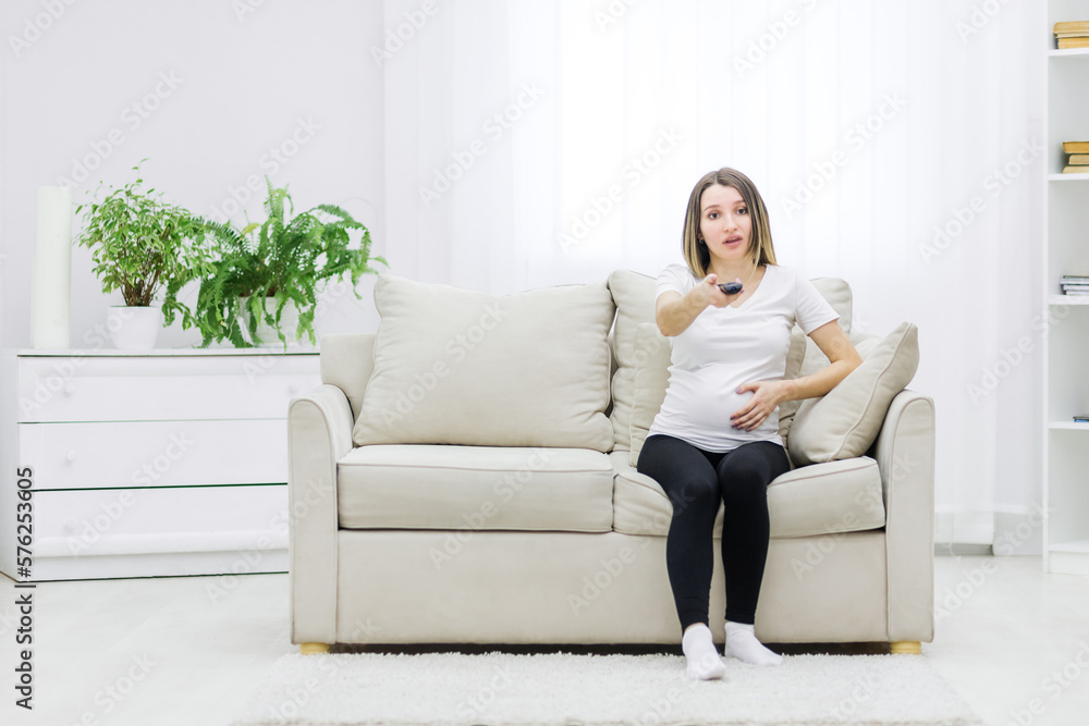 Pregnant woman facial expression sitting on sofa with remote control.