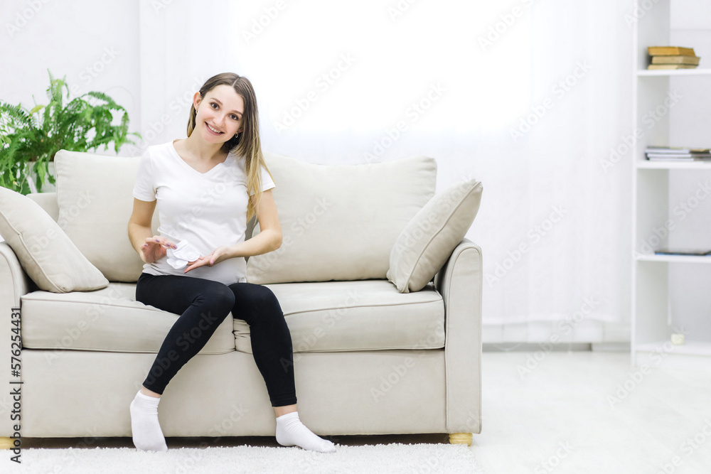 Smiling pregnant woman sitting on white sofa with two small socks.