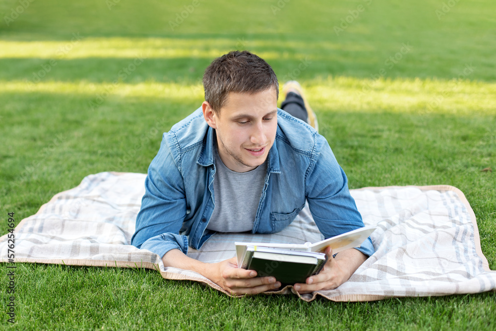 Young man relaxing and reading book while lying on lawn. Happy Man Lying On Green Grass and study remotely. Adult student reading in the park. Enjoying student life. smiling man working on outdoor Young man relaxing and reading book while lying on lawn. Happy Man Lying On Green Grass and study remotely. Adult student reading in the park. Enjoying student life. smiling man working on outdoor