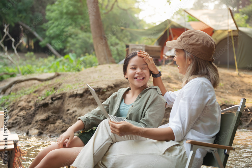 Pleased happy mother and daughter reading a book and using laptop while relaxing on the deck chairs in the river, sit near a camp and tent, drink coffee in a pine forest. Camping, recreation, hiking. Pleased happy mother and daughter reading a book and using laptop while relaxing on the deck chairs in the river, sit near a camp and tent, drink coffee in a pine forest. Camping, recreation, hiking.