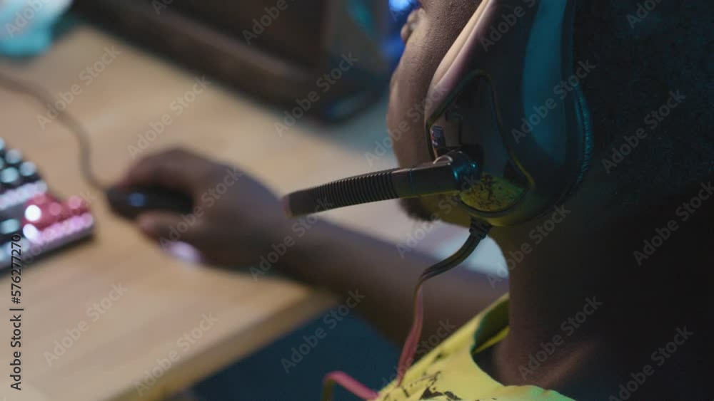 Over-the-shoulder shot of anonymous young African American gamer sitting at desk while playing online videogame with keyboard and mouse, adjusting microphone on headset and talking Over-the-shoulder shot of anonymous young African American gamer sitting at desk while playing online videogame with keyboard and mouse, adjusting microphone on headset and talking