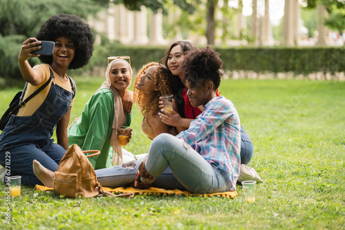 Group of smiling multiracial female best friends sitting together on blanket with fruits enjoying at picnic in the park - group of healthy friends having a picnic #576278229