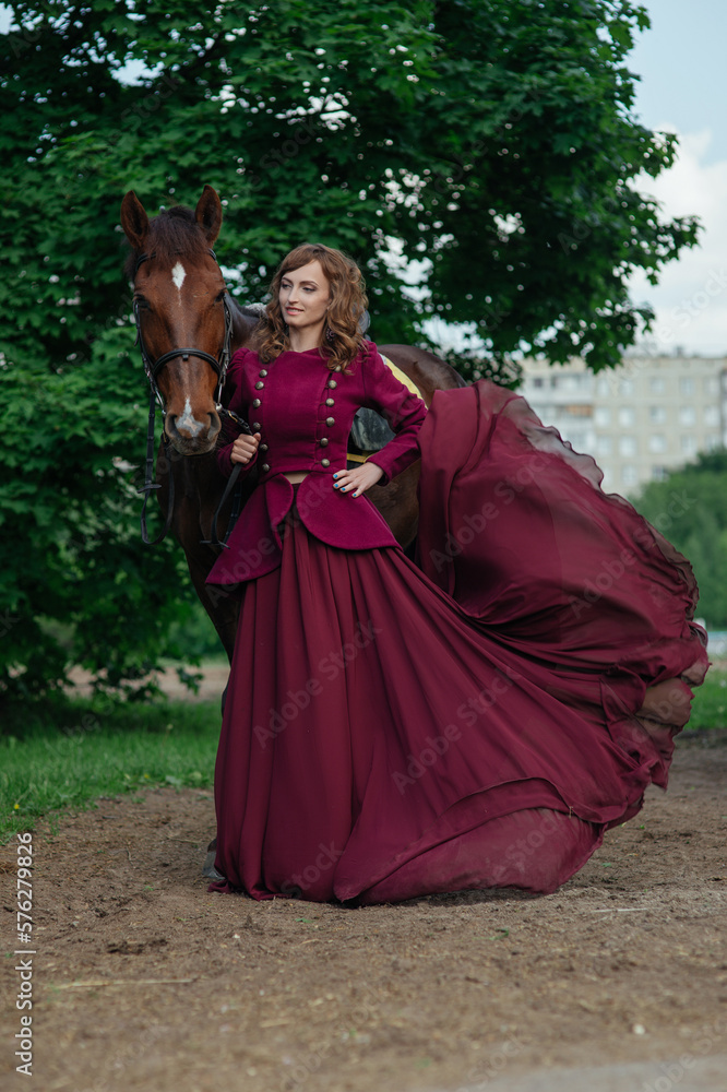 Beautiful elegant woman in long flowing dress and thoroughbred brown horse. Walk with horse in woods. Aristocratic young lady holding long vintage dress and well-bred stallion. Traditional horse farm