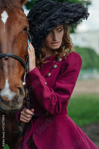 Old fashioned portrait of young woman in vintage purple suit and black hat is standing next to a horse. Elegant aristocratic young lady holding a horse by the reins. Horses and people. Animal friends © Anastasiia