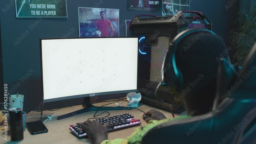 Over-the-shoulder shot of African American teenager with headset sitting at home in front of gaming computer with empty white screen and playing. Suitable as mockup, chromakey template