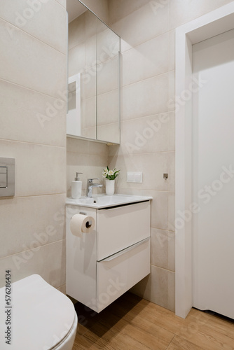 Interior design of aesthetics bathroom interior with white washbasin, big mirror, silver tap, beige tiles, vase with flowers, wooden panels floor and personal accessories. Home decor. Template.