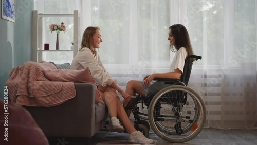Supportive woman holds hands of girlfriend with disability in wheelchair after accident trying to cheer up. Lesbian couple enjoys time together at home
