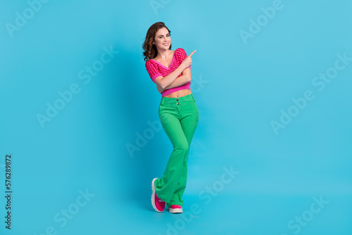Full size photo of cheerful optimistic woman with wavy hairstyle wear pink top directing empty space isolated on blue color background