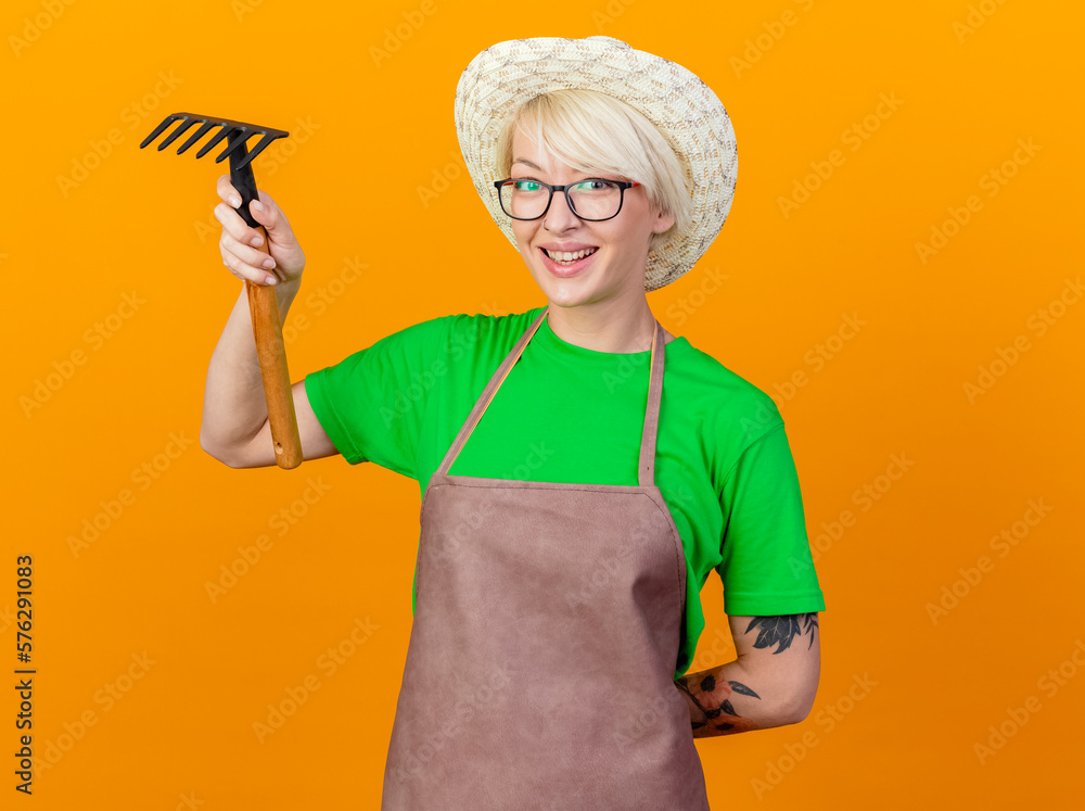 young gardener woman with short hair in apron and hat holding mini rake looking at camera smiling cheerfully standing over orange background young gardener woman with short hair in apron and hat holding mini rake looking at camera smiling cheerfully standing over orange background