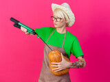 young gardener woman with short hair in apron and hat holding pumpkin cutting it with hedge clippers standing over pink background
