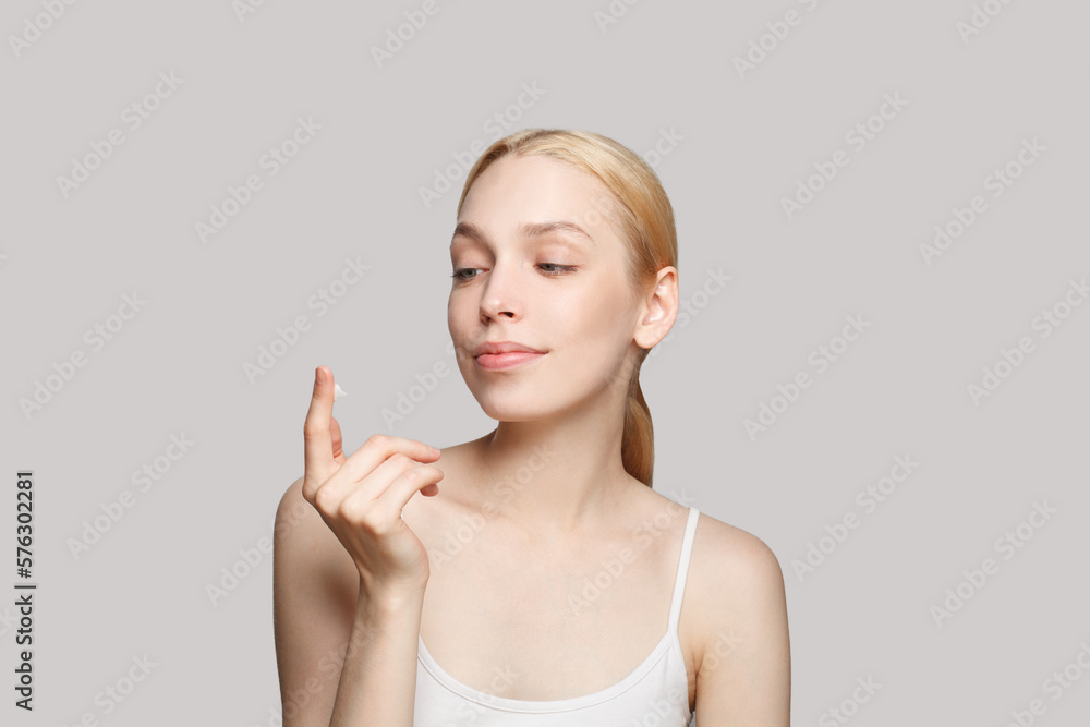 Portrait of pretty woman with blonde ponytail smiling and holding white moisturizing cream on her finger isolated on grey Portrait of pretty woman with blonde ponytail smiling and holding white moisturizing cream on her finger isolated on grey