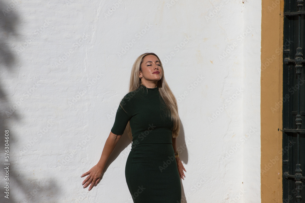 Beautiful blonde woman with straight hair dressed in green dress. The woman leaning on white wall looks up with her eyes closed as the sun rays enter.