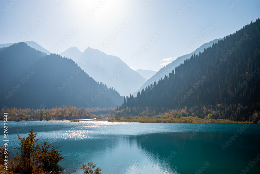 Autumn landscape. Tall old firs grow on the slopes of the mountains surrounding the mountain lake. A beautiful view of high mountains in a blue haze and a lake with azure water.