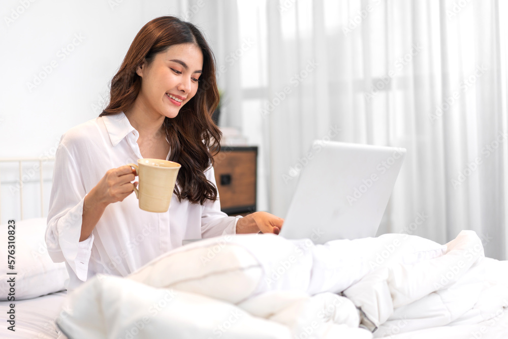 Young smiling happy beautiful asian woman relaxing using laptop computer in the bedroom at home.Young creative girl working and typing on keyboard.work from home concept Young smiling happy beautiful asian woman relaxing using laptop computer in the bedroom at home.Young creative girl working and typing on keyboard.work from home concept