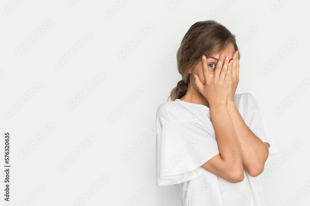 Young pretty blonde woman peeking through her fingers isolated over white background with copy space. Shy teen girl covering face with hands. Young pretty blonde woman peeking through her fingers isolated over white background with copy space. Shy teen girl covering face with hands.