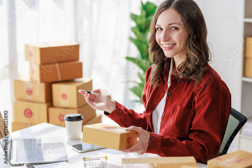 Concept of parcel delivery and selling online,Female seller or retailer write the customer's address on the parcel box to prepare for delivery and using laptop checking day's sales on the online store