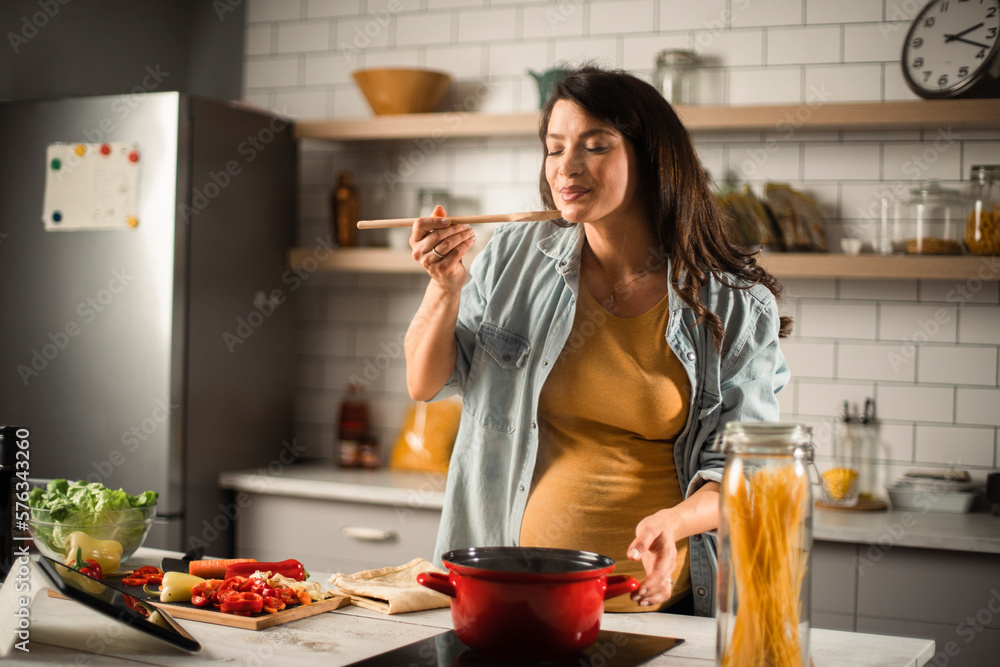 Beautiful pregnant woman preparing delicious food. Smiling woman cooking pasta at home.