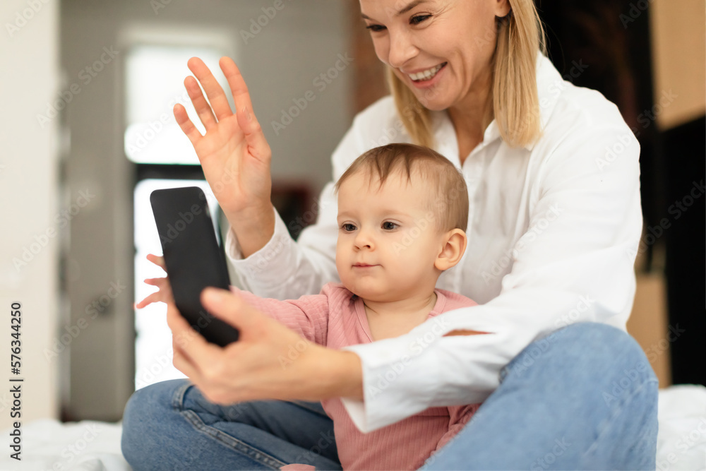 Smiling mother and her little daughter having video call on smartphone, sitting on bed and waving at phone screen Smiling mother and her little daughter having video call on smartphone, sitting on bed and waving at phone screen