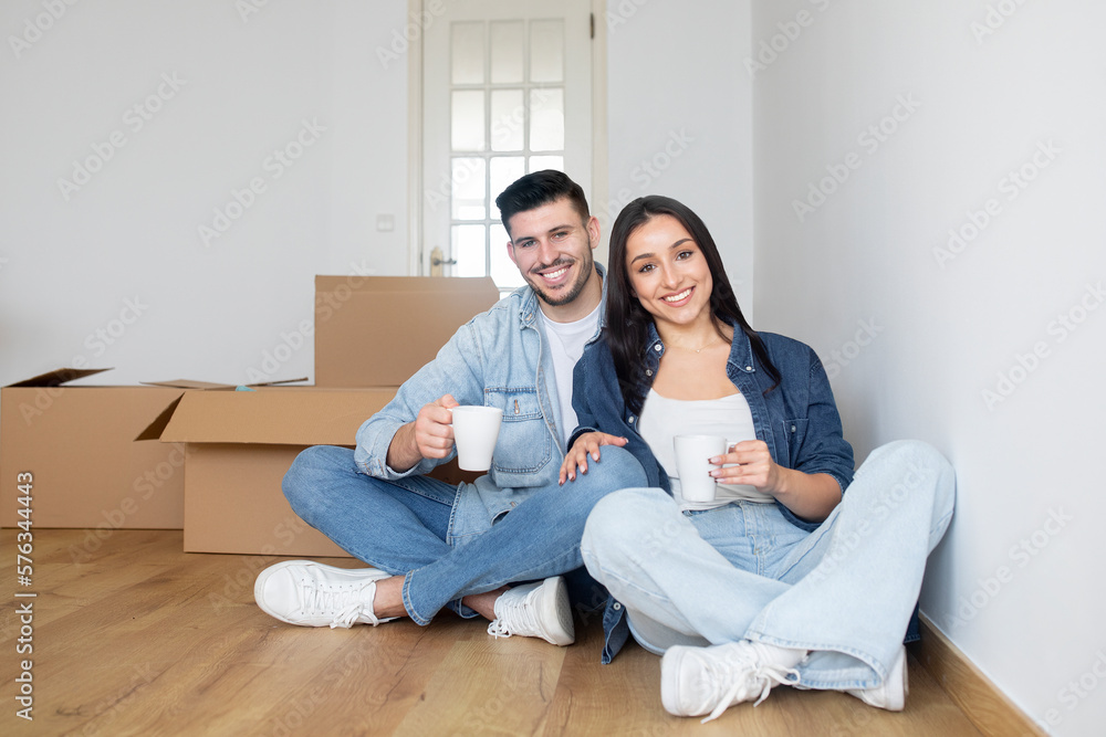 Happy young couple moving home, sitting among cardboard boxes and drinking coffee Happy young couple moving home, sitting among cardboard boxes and drinking coffee