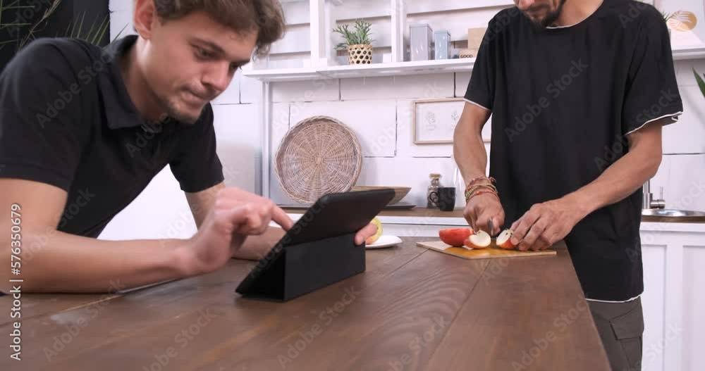 Two young men are using a digital tablet in the kitchen. Have fun spending free time together. One is looking at the screen while the other cuts an apple, close up Two young men are using a digital tablet in the kitchen. Have fun spending free time together. One is looking at the screen while the other cuts an apple, close up